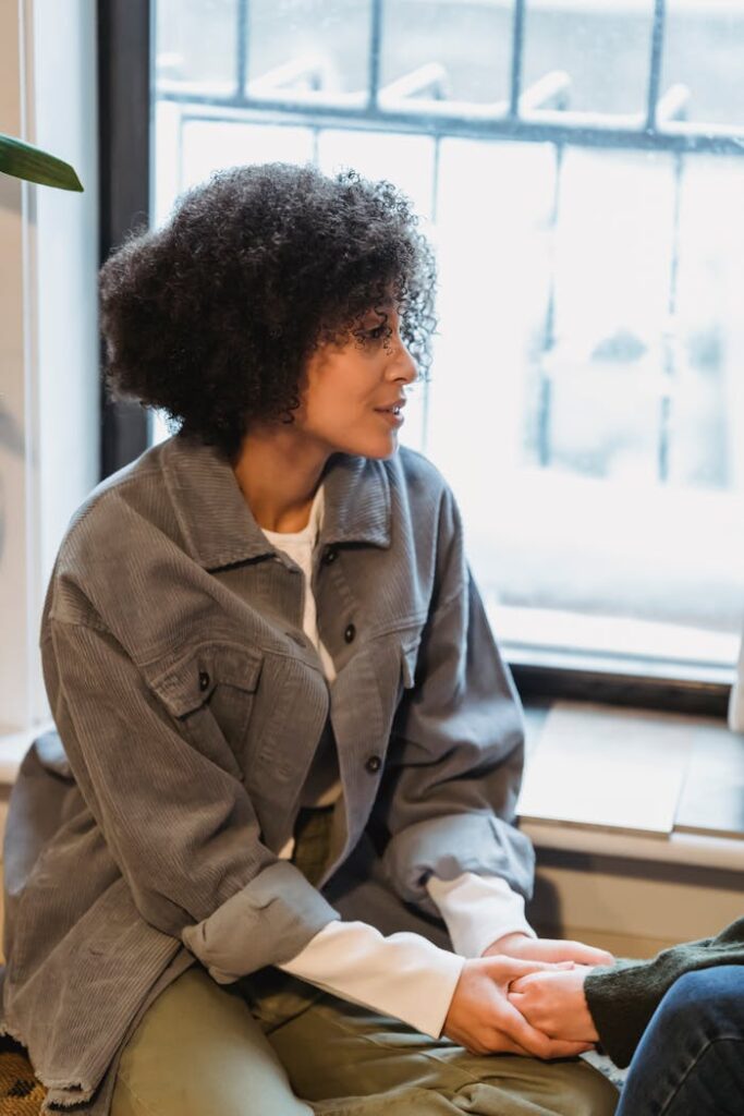 Serious young African American woman with dark curly hair in casual clothes holding hand of crop unrecognizable friend sitting near window at home