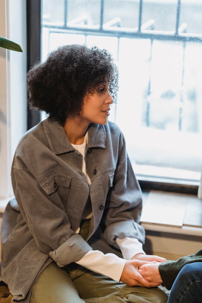 Serious young African American woman with dark curly hair in casual clothes holding hand of crop unrecognizable friend sitting near window at home